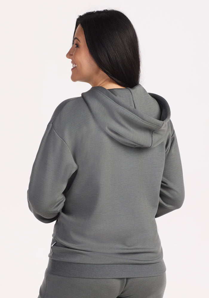 A woman with long dark hair, smiling and standing with her back to the camera, wears the Woolx Avery Hoodie in Sedona Sage against a plain light background.