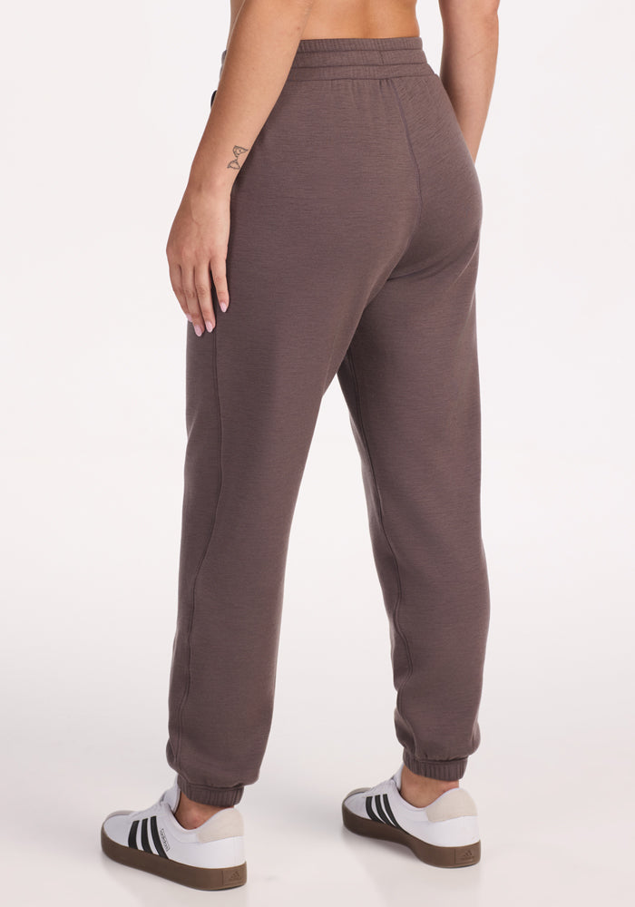 A person wearing Woolx Parker Sweatpants in Plum Truffle stands with their back to the camera against a plain white background, paired with white sneakers featuring black stripes.