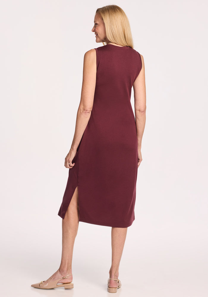A woman with long blonde hair wears the Woolx Cassie Dress in Toasted Garnet, a sleeveless, knee-length Merino wool tank dress with a side slit, paired with beige flats. She stands facing away from the camera against a plain white background.