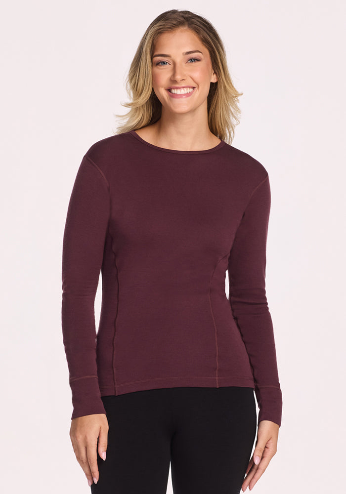 A woman with shoulder-length blonde hair smiles while wearing the Woolx Hannah Long Sleeve Top in Chocolate Fig and black pants, standing against a plain light background. 