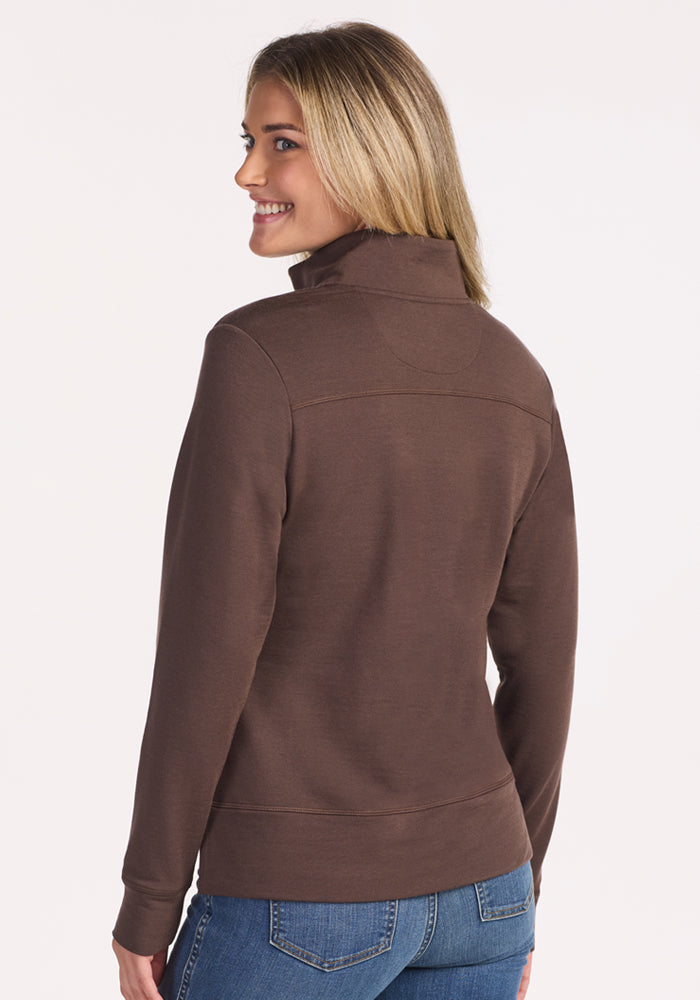A woman with blonde hair, smiling and standing slightly turned from the camera, wears the Woolx Annie 3 Button Pullover in Bracken with blue jeans against a plain, light background.