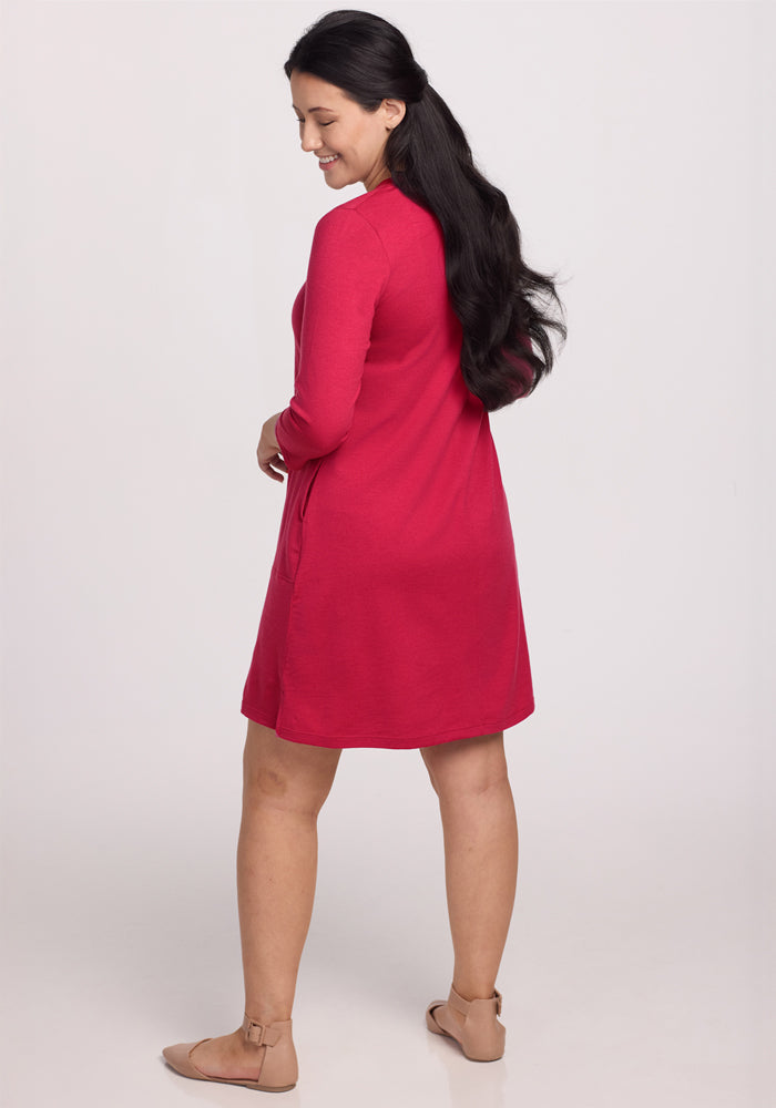 A woman with long dark hair, in the Woolx Rissa A Line Dress - Poppy Spark and tan flats, stands slightly turned against a white background—ideal for fall-winter layering.