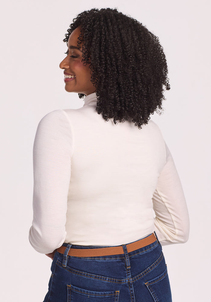 A woman with curly hair smiles, facing away from the camera, wearing the Woolx Kennedy Bodysuit in Buttercream with blue jeans and a brown belt against a plain light background—a true layering essential.