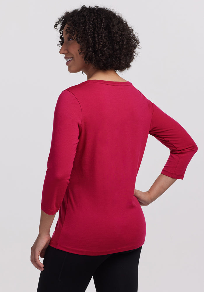 A person with short, curly hair stands with their back to the camera, wearing the Woolx Jenny 3/4 Sleeve Crew Neck in Poppy Spark and black pants against a plain, light-colored background.