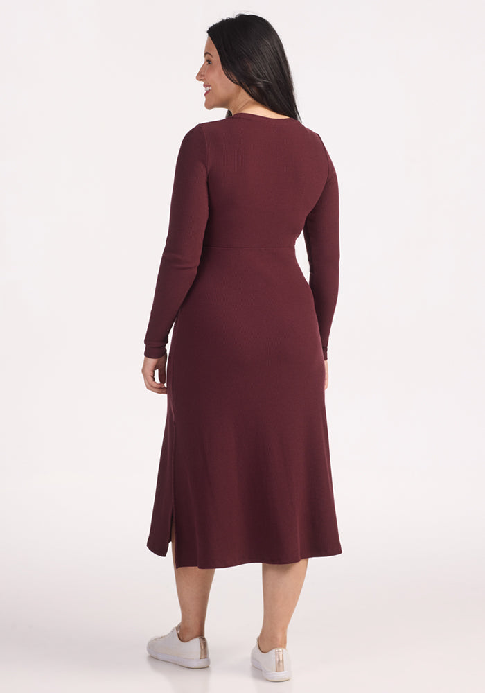 A woman with long dark hair, wearing the Woolx Caroline Ribbed Twirl Dress in Toasted Garnet and white sneakers, stands facing away from the camera against a plain white background.