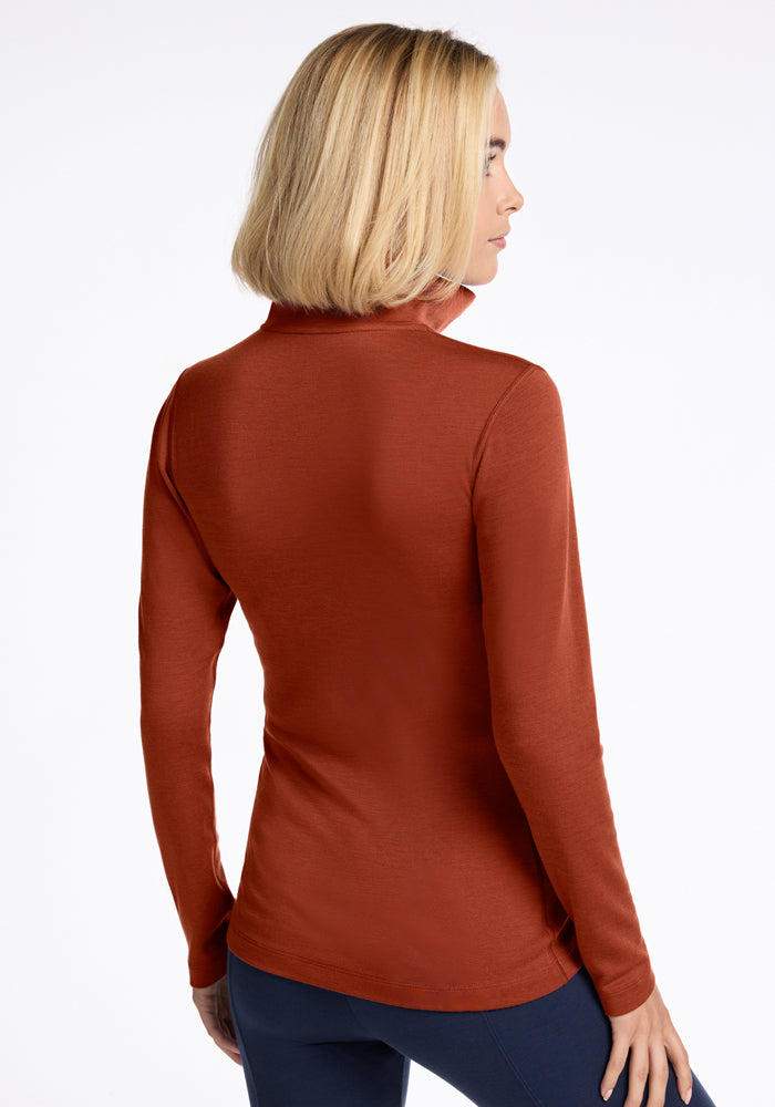 A woman with shoulder-length blonde hair is seen from behind, wearing the Woolx Brooke 1/4 Zip in Maple Leaf and dark pants against a plain white background.