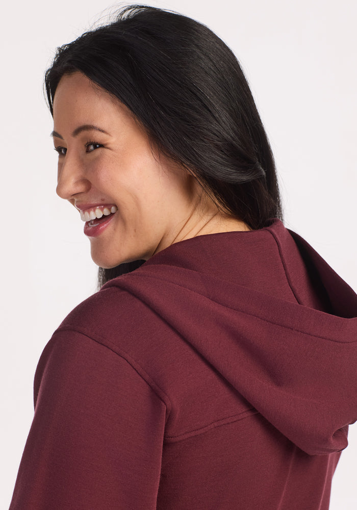 A woman with long dark hair smiles over her shoulder in the Woolx Darby Zip Up Hoodie in Toasted Garnet, facing slightly away from the camera. The cozy style stands out against a crisp white background.