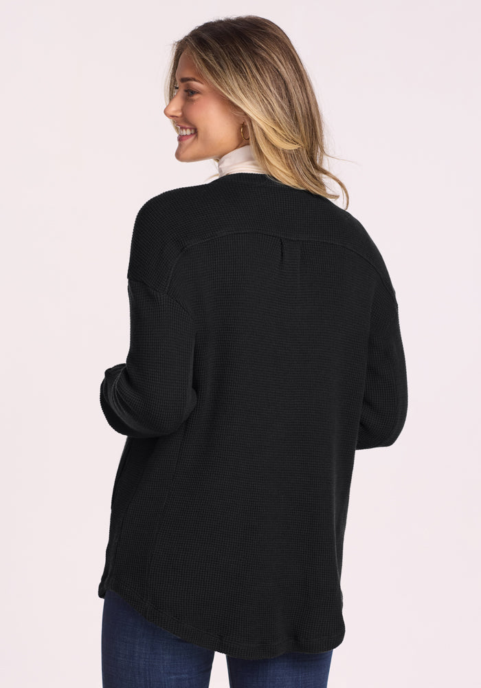 A woman with long, light brown hair smiles over her shoulder, wearing the Woolx Oaklynn Waffle Shacket in black over a white turtleneck and blue jeans. The background is plain and light-colored.