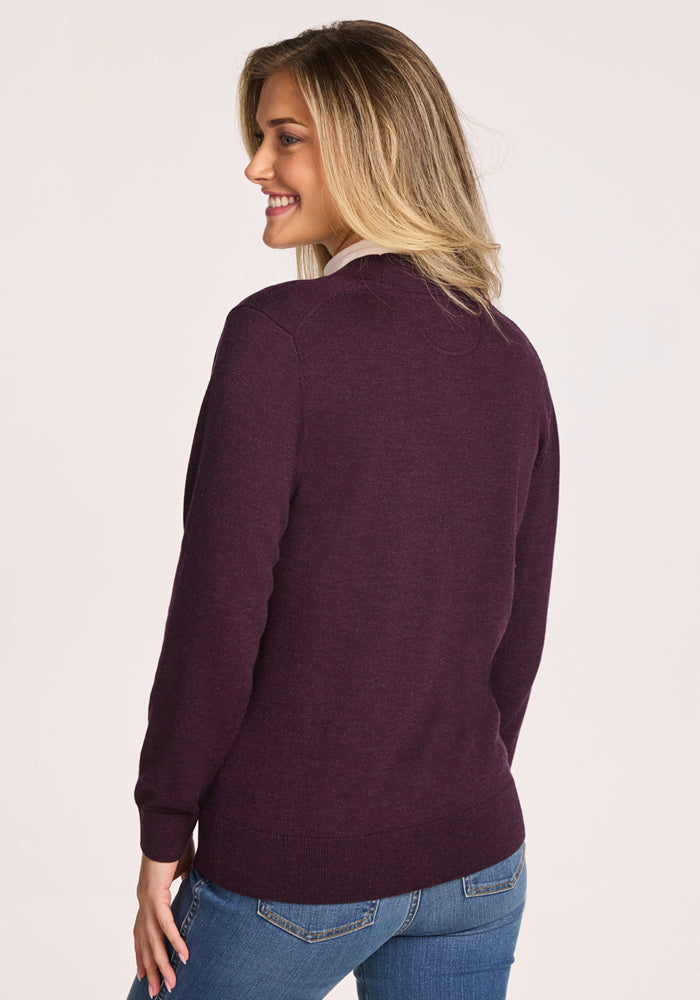 A blonde woman with a bright smile stands looking over her shoulder, wearing the Woolx Gia Sweater in Deep Plum and blue jeans, set against a light background.