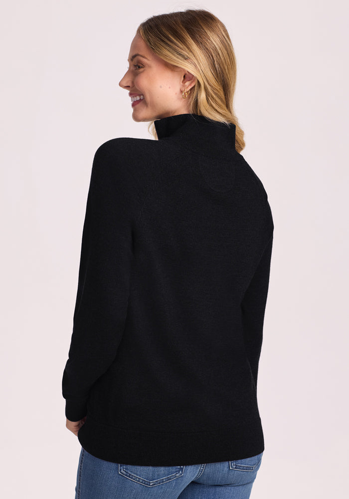 A woman with blond hair stands facing away, smiling to the side. She wears Woolx's Collins Half Zip Sweater in black and blue jeans against a light background.