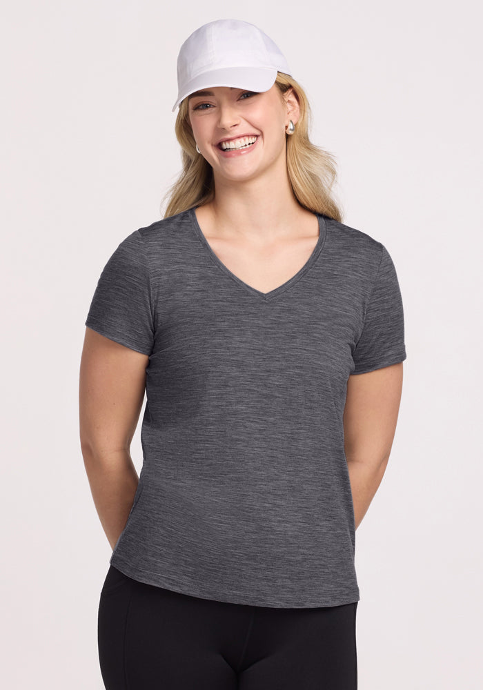 A woman stands smiling with her hands behind her back, wearing the Woolx Junie V Neck in Cinder Melange, a white cap, and black pants against a plain light background. 