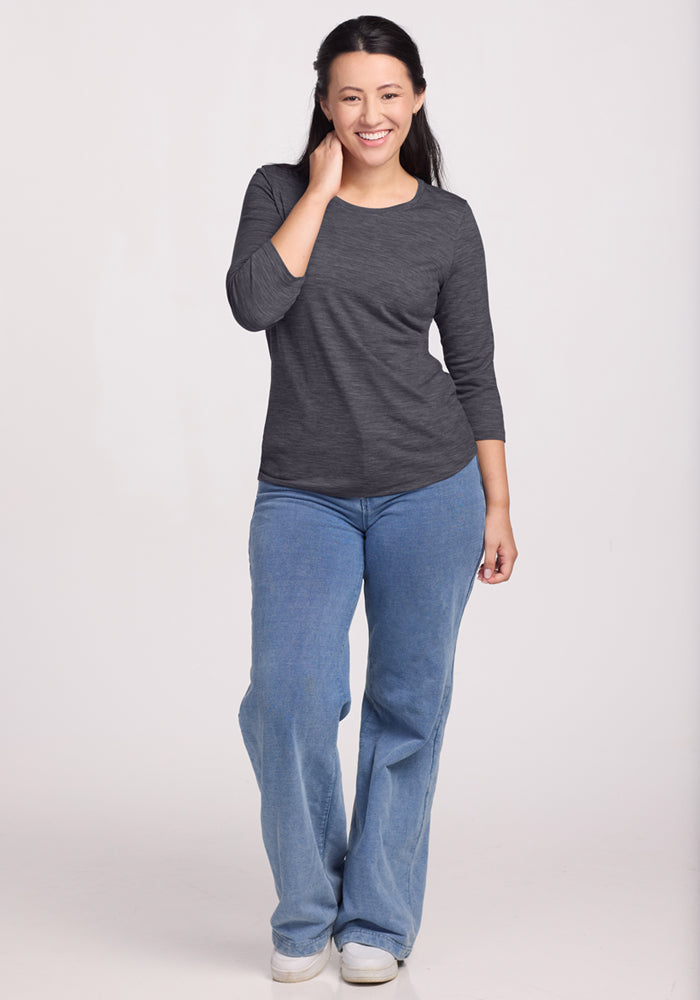 A woman with long dark hair, wearing the Woolx Jenny 3/4 Sleeve Crew Neck in Cinder Melange and light blue jeans, stands smiling with one hand touching her hair against a plain light background.