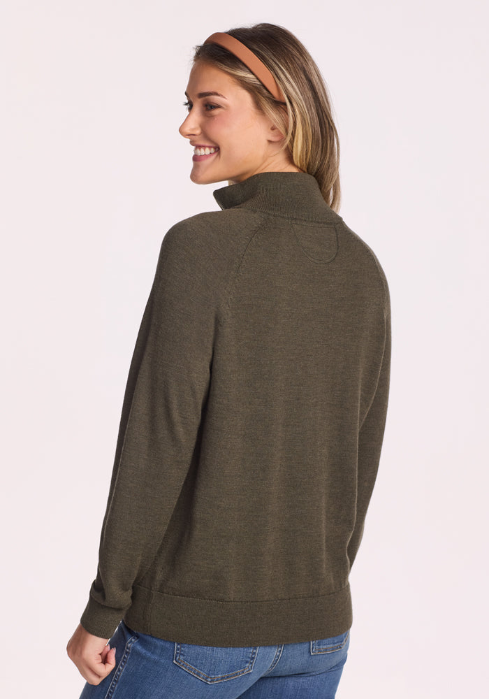A woman with long hair, smiling and facing away from the camera against a light background, wears the Woolx Collins Half Zip Sweater in Pine Needle and blue jeans, accessorized with a brown headband.