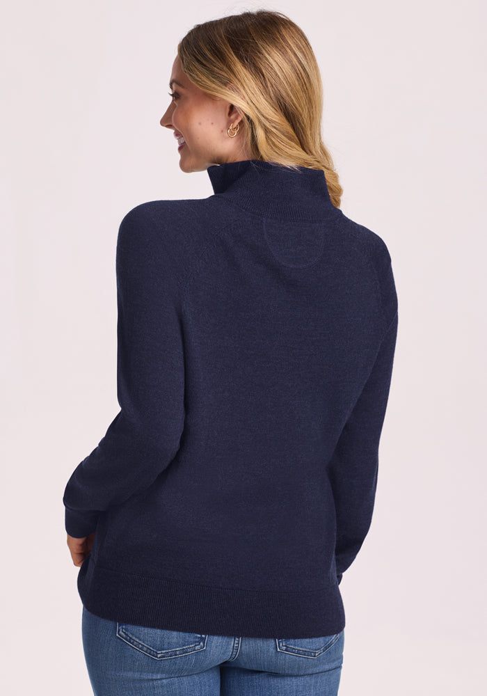 A woman with long blonde hair models the Woolx Collins Half Zip Sweater in Storm Cloud and blue jeans, standing with her back to the camera and smiling over her shoulder against a plain light background.