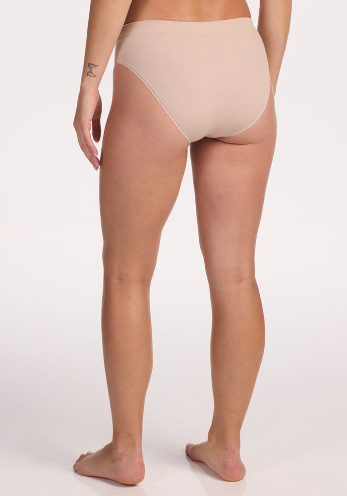 A person stands barefoot on a white background, shown from the back, wearing Woolx Maeve Bikini in Sand. A small tattoo is visible on the left wrist.