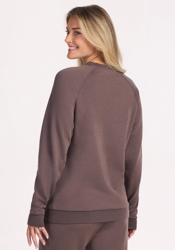 A woman with long, blonde hair smiles over her shoulder, wearing the Woolx Bailey Crewneck Sweatshirt in Plum Truffle with matching pants, standing against a plain light background.