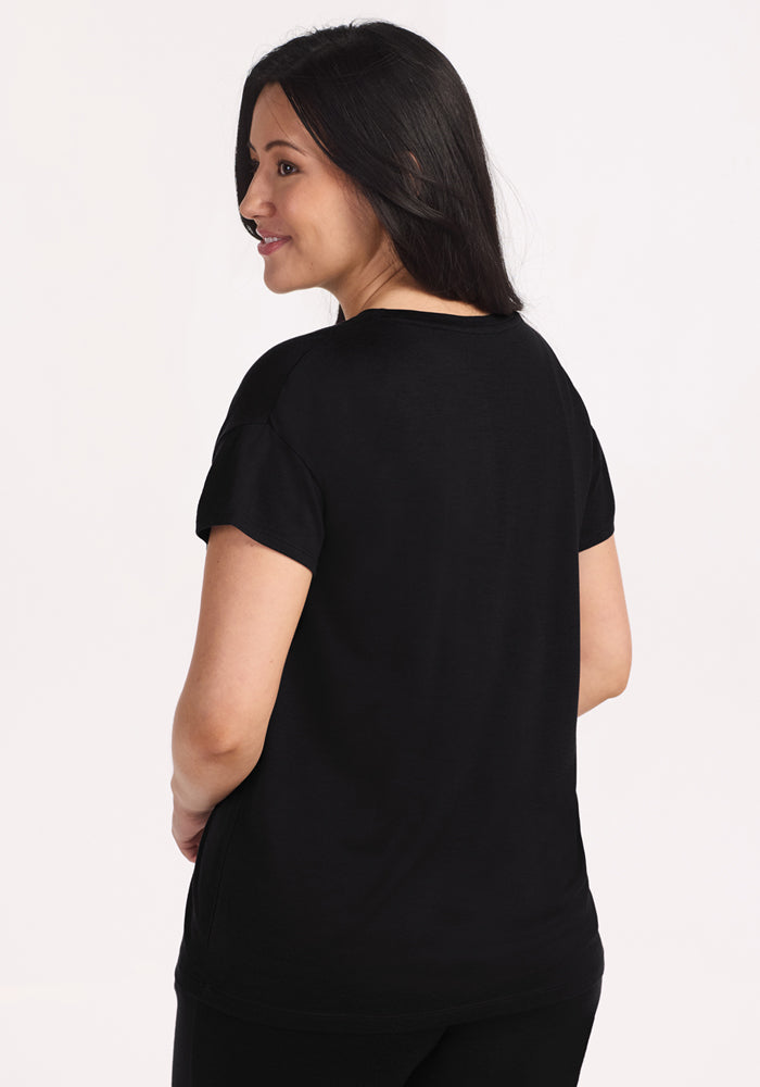 A woman with long dark hair, wearing the Woolx Billie Lounge Tee in black and matching pants, stands turned slightly away from the camera, smiling over her shoulder against a plain light background.