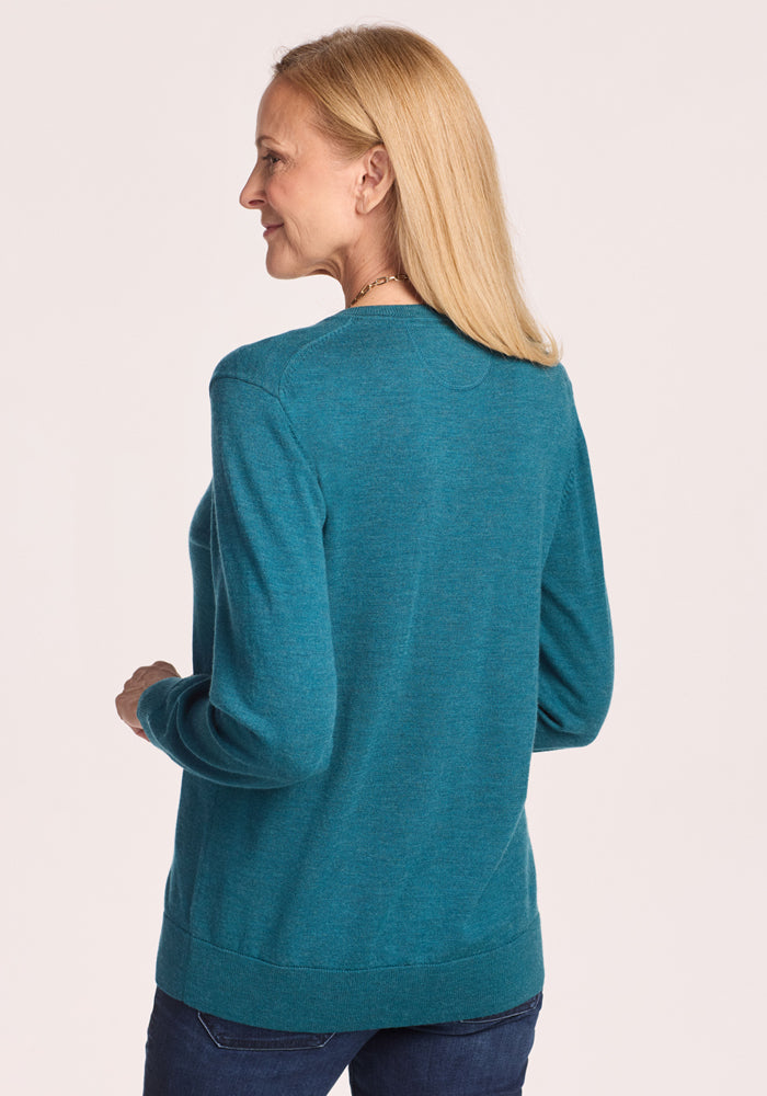 A woman with straight blonde hair, wearing the Woolx Francesca Sweater in Harbor Teal and dark jeans, stands facing away and looks to her left against a light background.