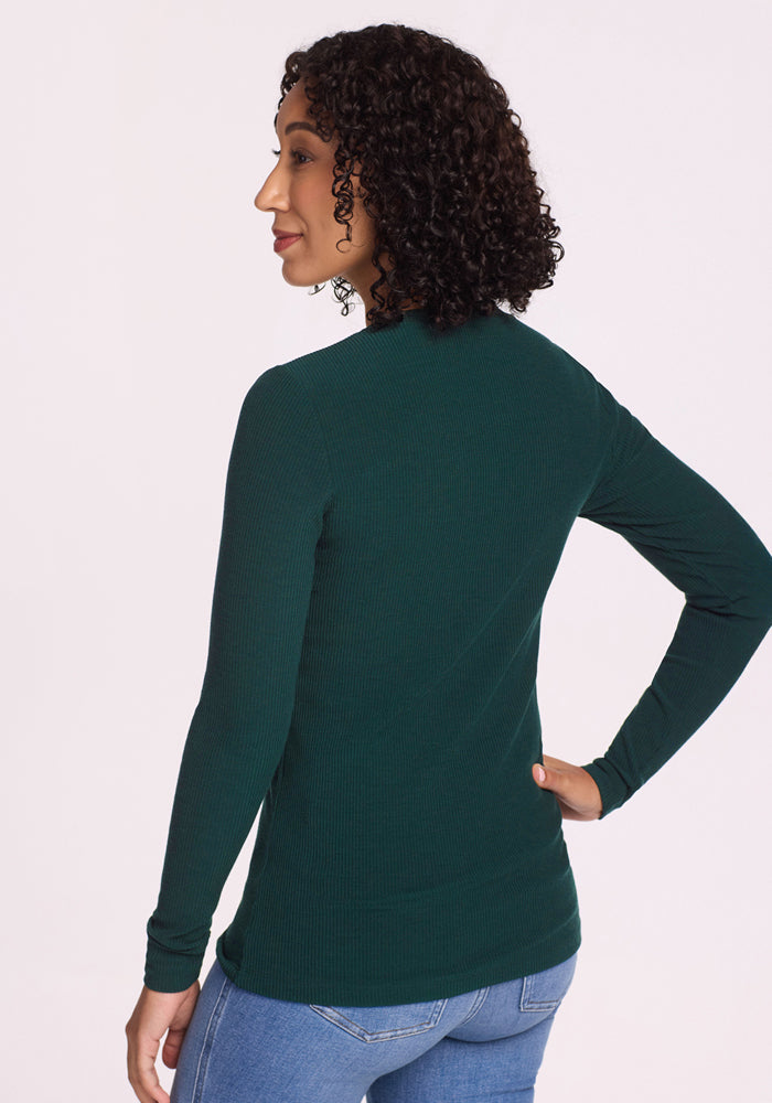 A woman with curly dark hair stands facing away, wearing the Woolx Hadley Ribbed Crew in Deep Timber and light blue jeans. She has one hand on her hip, set against a plain white background.