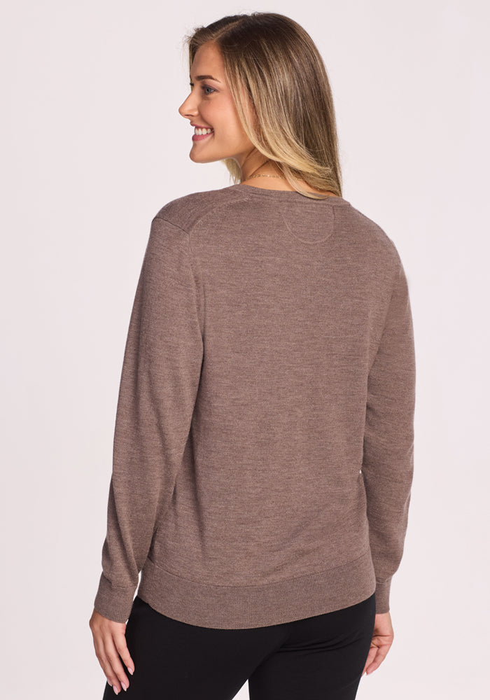 A woman with long, light brown hair smiles while modeling the Woolx Francesca Sweater in Simply Taupe and black pants. She’s turned slightly to the side, displaying the back and side of the sweater against a plain light background.