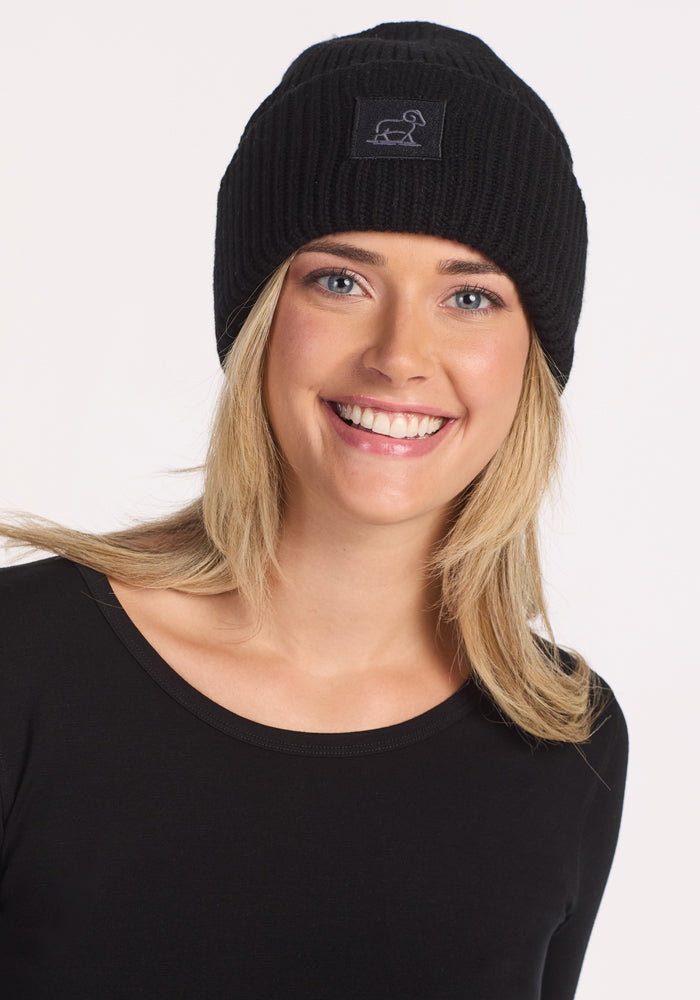 A smiling blonde woman models the Woolx Ria Chunky Beanie in black with a matching top, standing against a plain white background—an ideal way to highlight this cozy winter accessory.