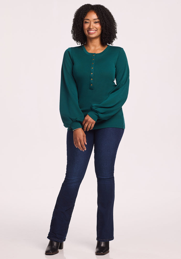 A woman with curly hair smiles while wearing the Woolx Charlotte Puff Sleeve Top in Riverbank Green, paired with dark blue jeans and black boots, standing against a plain light background.