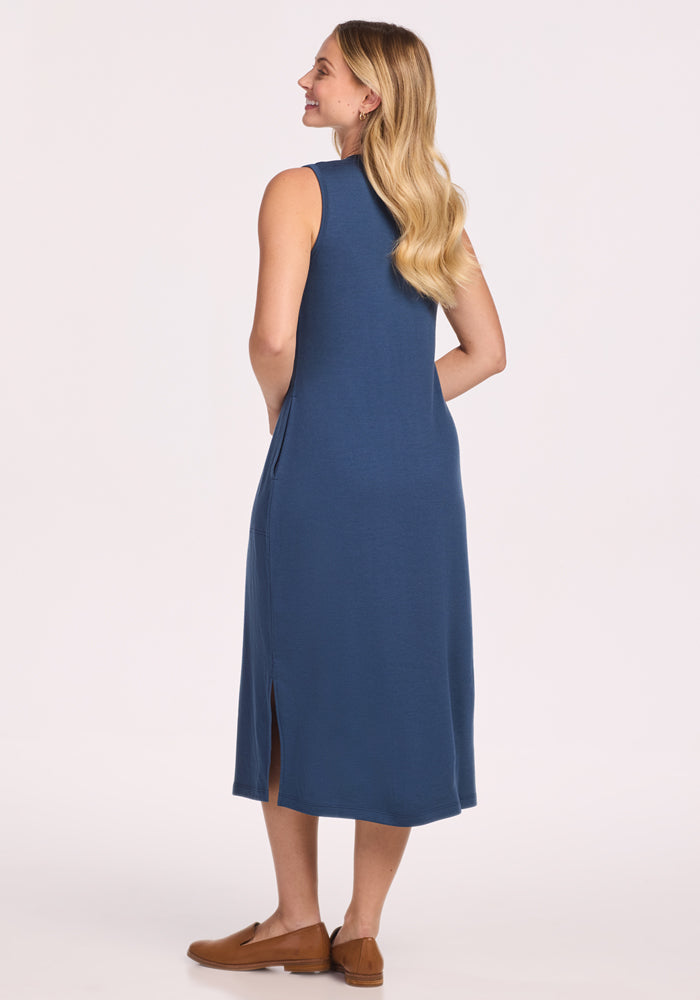 A woman with long, wavy hair smiles, wearing the Woolx Cassie Dress - Starry Night—a sleeveless, calf-length blue Merino wool dress with side slits and brown flats. Stylish and comfy on a plain light background, it's the perfect travel dress.