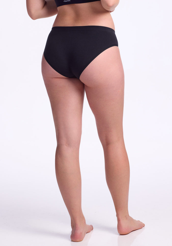A woman stands barefoot on a white background, facing away from the camera. She wears Woolx Maeve Bikini - Black merino wool underwear and a matching bra, with her arms relaxed by her sides and legs slightly apart.