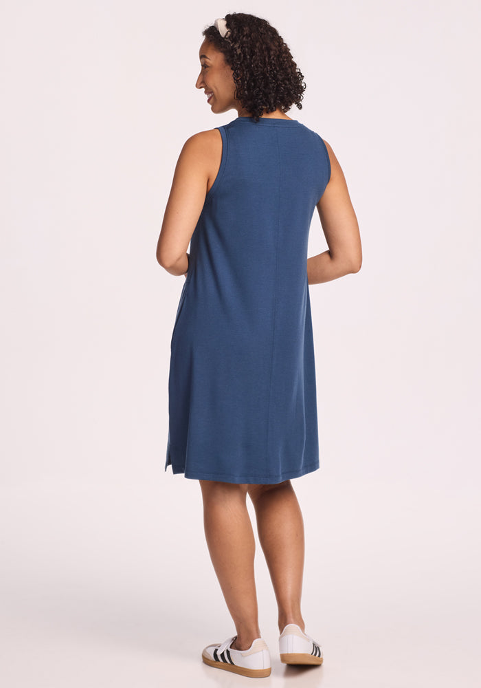 A woman with curly hair stands smiling with her back to the camera against a plain light background, wearing the Woolx Clara Dress - Starry Night and white sneakers with black stripes.