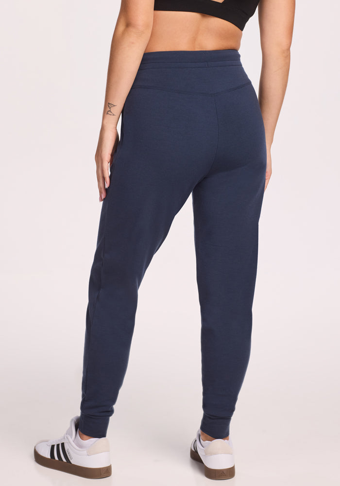 A person in a black crop top, Woolx Willa Joggers in Deep Navy, and white sneakers with black stripes stands facing away, displaying the back of the outfit against a plain light background.