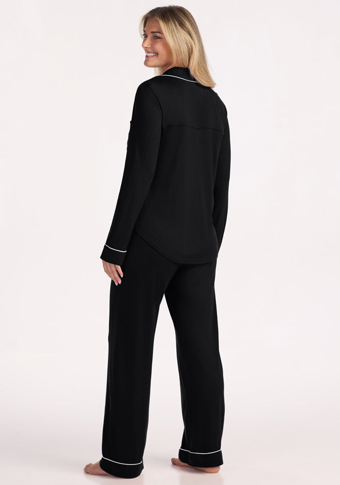 A blonde woman smiles while standing barefoot, facing away and turning her head back. She wears the Woolx Harper FeatherTouch® Pajama Set in black, featuring white piping on the collar, cuffs, and pant hems.