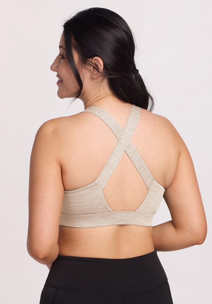 A woman with long dark hair smiles at the camera, showing the crisscross straps of the Woolx Livi Bra in Cream Heather, paired with black leggings against a plain, light background.