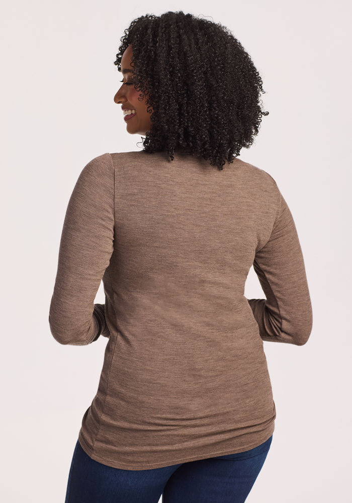 A woman with curly hair stands facing away from the camera, wearing the Woolx Sage Ribbed Turtleneck in Simply Taupe and blue jeans. Her arms are relaxed by her sides against a plain, light background.