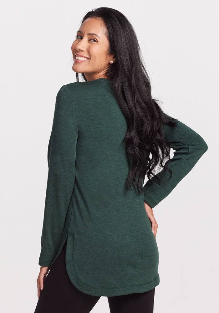 A woman with long dark hair smiles over her shoulder in the Woolx Blair Tunic - Forest and black pants, enjoying lightweight warmth and odor-resistant comfort against a plain light background.