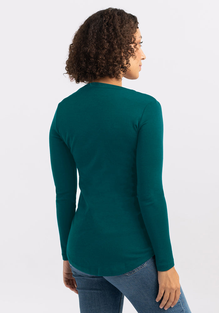 A person with short curly hair, wearing the Woolx Eva Tunic in Riverbank Green and blue jeans, stands facing away to display the back of the tunic against a plain light background.