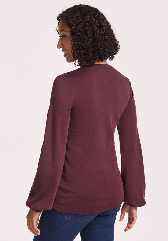 A woman with curly hair is seen from the back, smiling and wearing the Woolx Charlotte Puff Sleeve Top in Chocolate Fig with blue jeans, standing against a plain light background, one hand relaxed by her side.