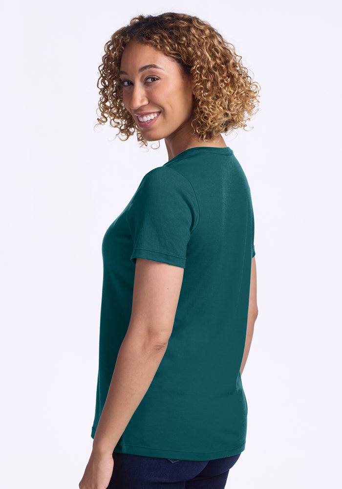 A woman with curly hair, wearing the Woolx Junie V Neck in Riverbank Green and dark pants, stands sideways and smiles at the camera against a plain light background.