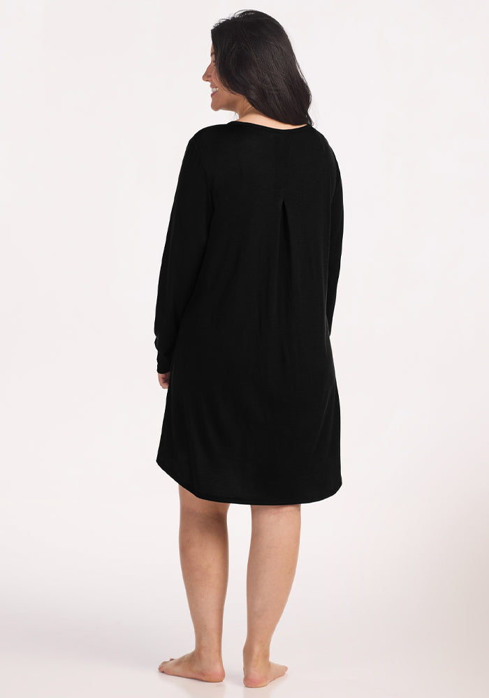A woman with long dark hair stands barefoot on a light background, smiling as she faces away from the camera in the Woolx Lucille Long Sleeve Nightgown - Black, enjoying the comfort of sustainable sleepwear.