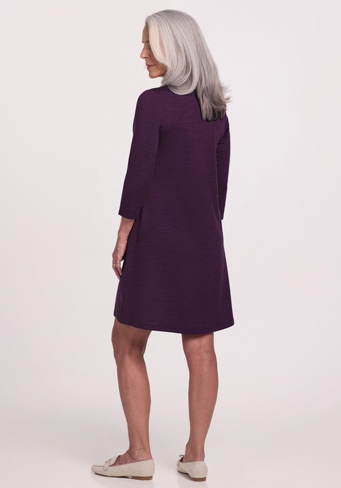 An older woman with long gray hair wears the Woolx Rissa A Line Dress in Deep Plum, a knee-length, long-sleeve Merino wool piece styled with beige flats, standing with her back to the camera against a plain light background—ideal for layering.