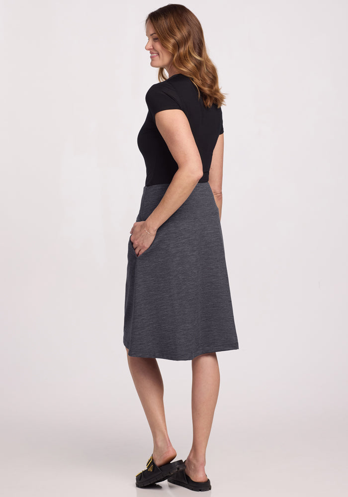 A woman stands sideways, smiling with her hand in the pocket of her Woolx Marilyn Flowy Midi Skirt in Cinder Melange. She wears a fitted black short-sleeve top and black slip-on shoes against a plain white background.
