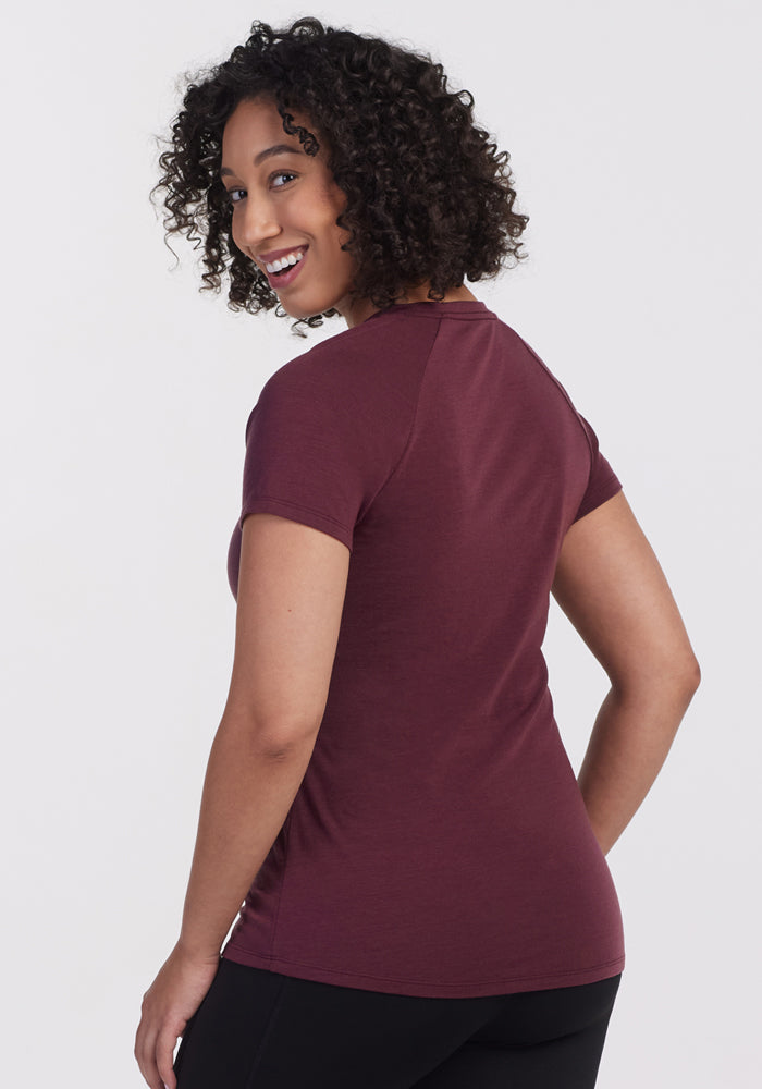 A woman with short curly hair smiles over her shoulder, wearing the Woolx Mia V Neck - Grape Harvest t-shirt and black pants, standing against a plain white background.