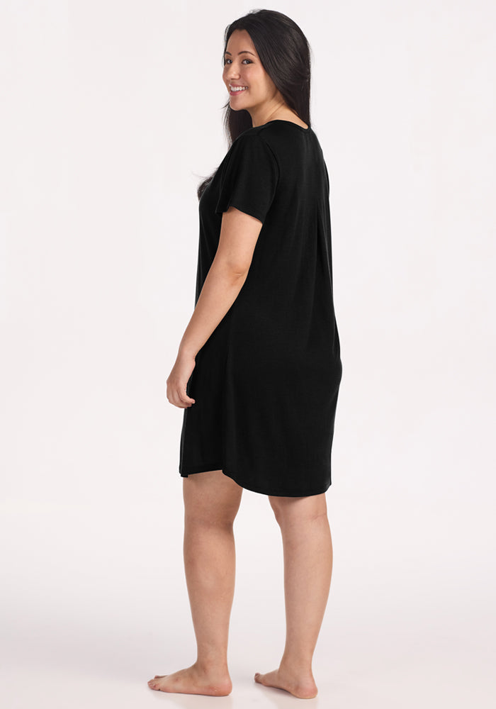 A woman with long dark hair stands barefoot, smiling over her shoulder in the Woolx Desi Nightgown - Black, a loose-fitting, short-sleeved Merino wool nightgown known for its temperature-regulating comfort against a plain light background.