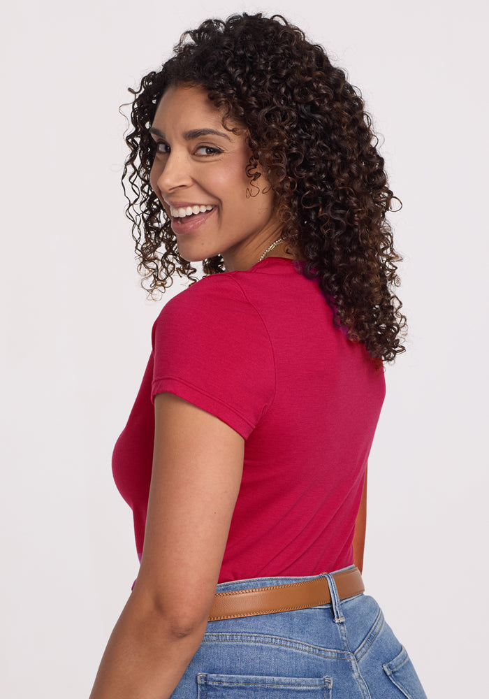 A woman with curly hair, wearing the Woolx Addie Crew Neck in Poppy Spark and blue jeans with a brown belt, smiles over her shoulder against a plain light background.