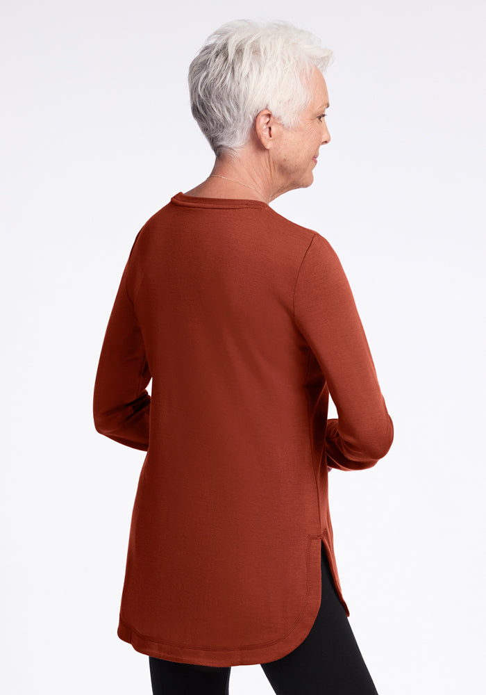 An older woman with short white hair is seen from the back, wearing the Woolx Blair Tunic in Maple Leaf paired with black pants, standing against a plain white background.