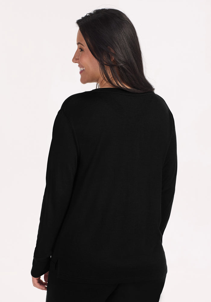 A woman with long dark hair wears the Woolx Ashlie Lounge Top in black, standing facing away and smiling over her shoulder. The plain, light background showcases the top's wrinkle-resistant, temperature-regulating Merino wool fabric.