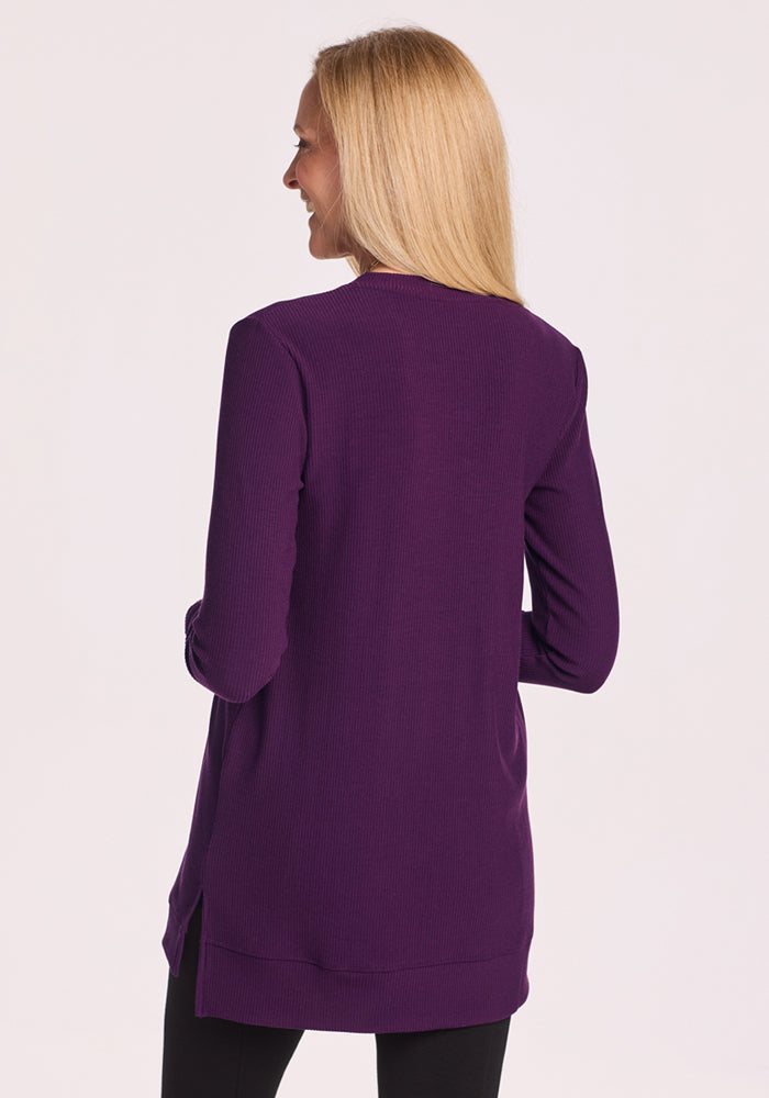 A woman with straight blonde hair stands facing away, wearing the Woolx Evie Ribbed Tunic in Majestic Purple with black pants, set against a light background.