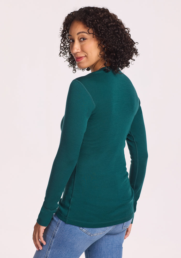 A woman with curly hair wears the Woolx Hannah Long Sleeve Top in Riverbank Green with blue jeans, standing turned slightly away and glancing over her shoulder, smiling against a plain light background.