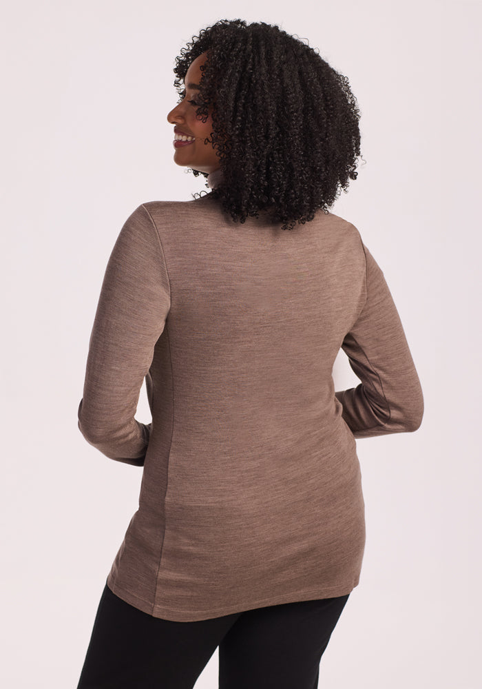 A woman with curly hair, wearing the Woolx Peyton Turtleneck in Simply Taupe and black pants, stands facing away from the camera against a plain light background.