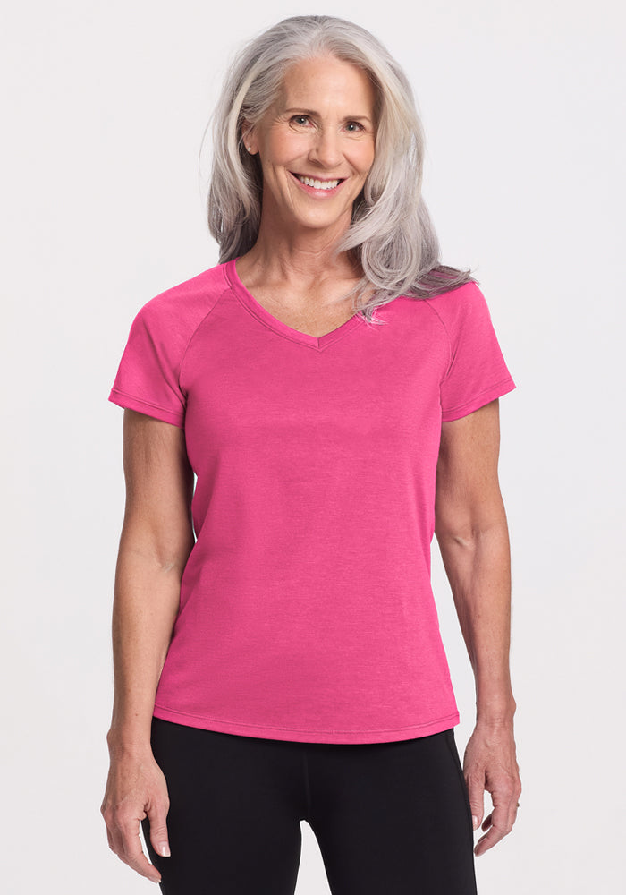 A smiling older woman with long gray hair wears the Woolx Mia V Neck in Raspberry Fizz and black pants, standing against a plain light background. 