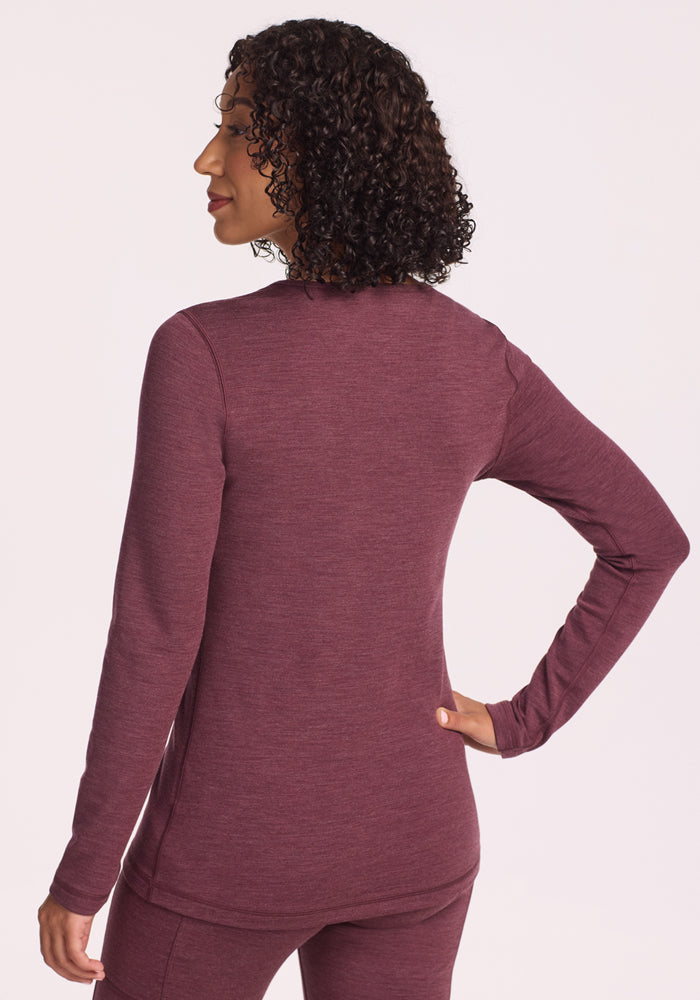 A person with curly hair stands with their back to the camera, wearing the Woolx Elsa Long Sleeve in Grape Harvest Melange. One hand rests on their hip, and the light background highlights the moisture-wicking merino wool top.